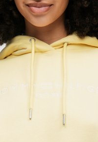 Κίτρινη φούτερ φούτερ φτιαγμένη από μαλακό ύφασμα με λευκό κεντημένο λογότυπο. Διαθέτει ασημένια μεταλλικά εξαρτήματα στις ρυθμιζόμενες κούμπες και χαλαρή εφαρμογή.