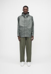 Ελαφρύ, γκρι μπουφάν με κουκούλα, φερμουάρ, μπροστινές τσέπες και ελαστικό κορδόνι. Συνδυασμένο με ελαιόχρεια παντελόνια και λευκά αθλητικά παπούτσια.