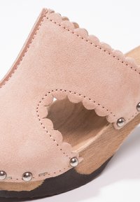 Sabots en daim rose avec un bord festonné, une base en bois et des accents de clous argentés. Les côtés ouverts améliorent la respirabilité et apportent une touche esthétique.