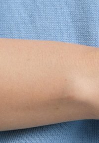 Lichtbeige arm met een gladde textuur, gedeeltelijk bedekt door een zachte, gebreide blauwe trui. De arm staat onder een lichte hoek, wat details en schaduw benadrukt.