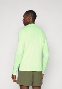 Maglietta sportiva a maniche lunghe di colore verde chiaro, realizzata in un tessuto traspirante, con un motivo testurizzato sulle maniche e un design con colletto alto.