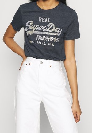 Frau trägt ein dunkelgraues SuperDry-T-Shirt mit weißem Text und Symbolen, kombiniert mit hoch taillierter weißer Hose, eine Hand in die Hüfte gestützt.