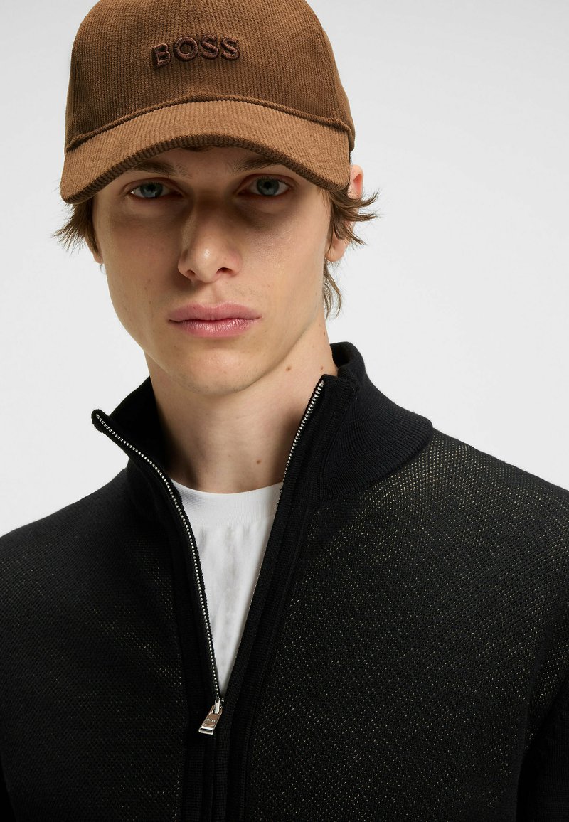 Brown cap with "BOSS" logo, black zip-up jacket with mesh texture, and white t-shirt underneath. Close-up of face and upper body.