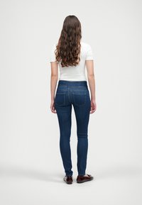 Pielāgotas tumši zilas džinsu bikses ar vidēja augstuma jostu, izteiksmīgu šuvju un aizmugurējām kabatām, kombinējot ar piegulīgu baltu krekla un tumšām kurpēm.