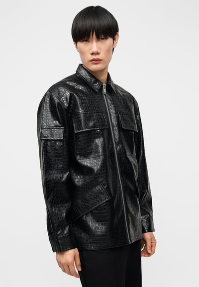 Νεαρός άνδρας με ίσια μαύρα μαλλιά που φοράει μαύρο δερμάτινο τζάκετ με φερμουάρ και υφή κροκόδειλου, με μεγάλα τσέπες με καπάκι.