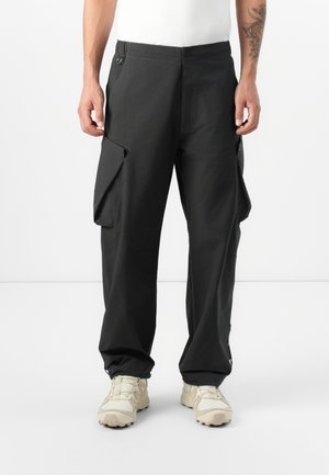 A person stands wearing black cargo pants with side pockets and light-colored sneakers, showcasing a casual, modern style against a plain background.