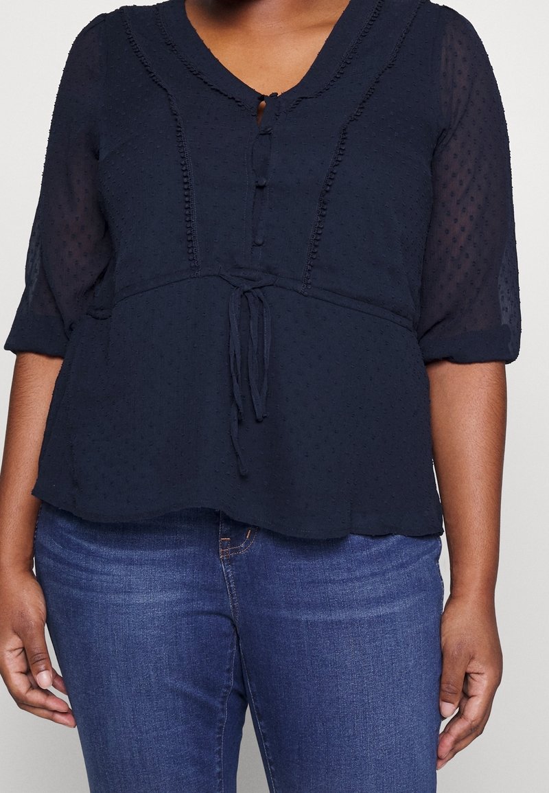 Marineblaue Bluse aus strukturiertem Stoff, mit Knopfleiste und Drei-Viertel-Ärmeln. Sie hat einen Bindegürtel an der Taille und einen gewellten Ausschnitt.