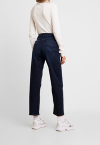 Tmavě modré džínové kalhoty s vysokým pasem a rovnými nohavicemi, s předními i zadními kapsami a jemnými švy. Spárováno s bílými teniskami.