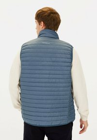 Blau gepolsterte Weste mit horizontaler Steppung, hohem Kragen und Logo auf der Rückseite. Über einem beigen Strickpullover mit geradem Schnitt getragen.
