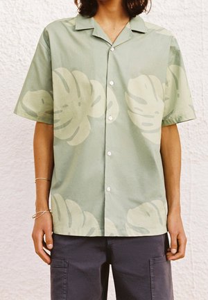 Košile s krátkým rukávem na knoflíky s velkými světle zelenými tropickými listy, nošená s tmavými cargo šortkami a tenkými náramky na levém zápěstí.