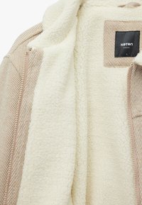 Beige cordjakke med en blød cremefarvet fleeceforing. Har en lynlåslukning og et mærke, der fremviser en blanding af teksturer og farver.