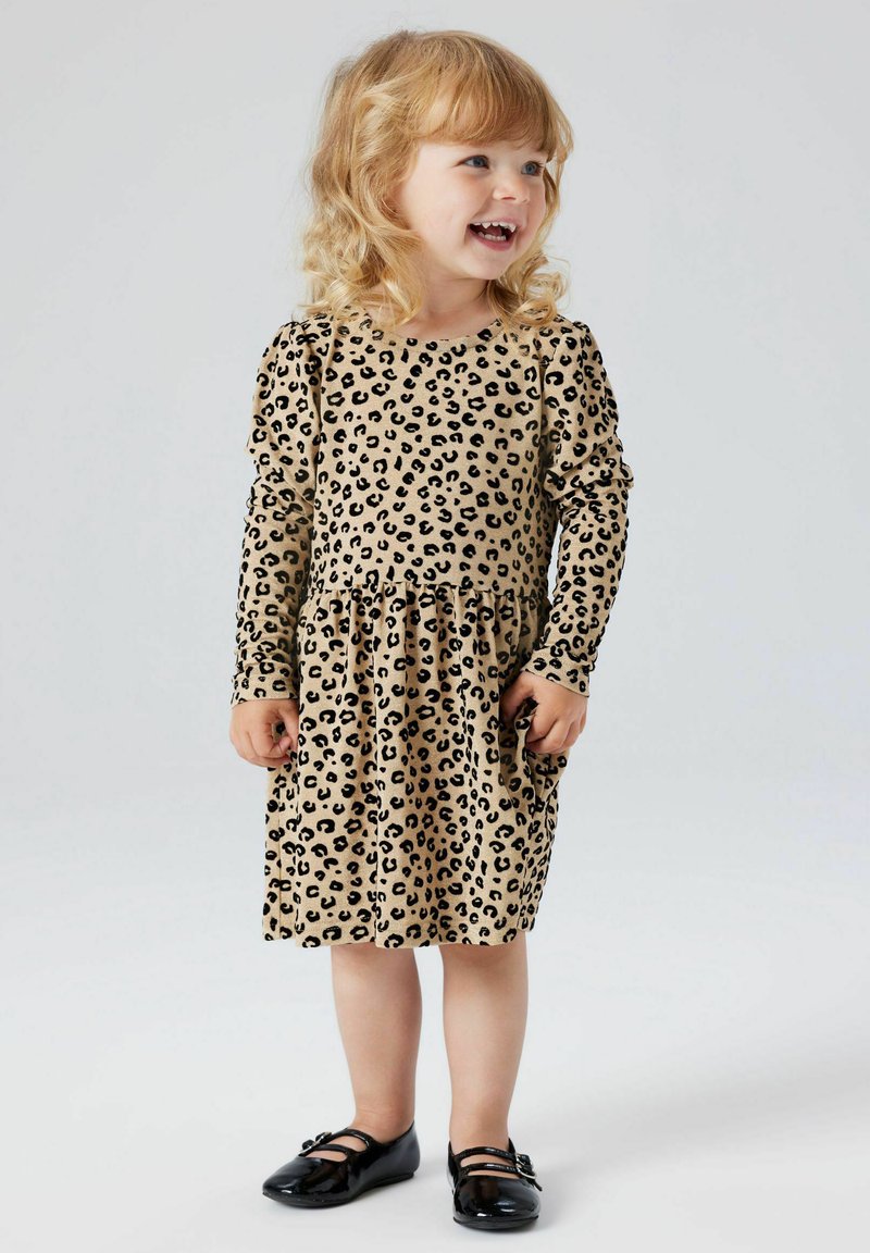 Bežs leopardu raksts kleita ar garām piedurknēm un savilkta josta. Gluda auduma un melni akcenti. Bērns valkā melnas Mary Jane kurpes.