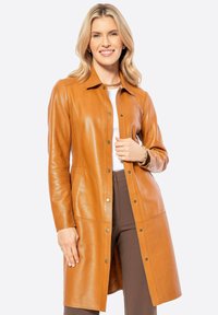 Tan lederen lange jas met een kraag, drukknoopsluiting, zijzakken en een gladde afwerking, gedragen over een wit topje en bruine broek.