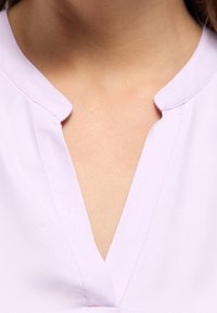 Světlá levandulová halenka s výstřihem do V a jemnou, hladkou texturou. Vyznačuje se minimalistickým designem bez ozdob.