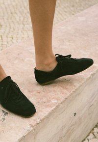 Scarpe in suede nere con punta arrotondata e design con lacci, caratterizzate da un profilo basso e una texture liscia, poggiate su una superficie di colore chiaro.
