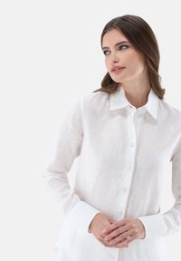 Camicia bianca in lino con bottoni frontali, collo a punta e maniche lunghe. Presenta un tessuto leggero e texturizzato e polsini a contrasto.