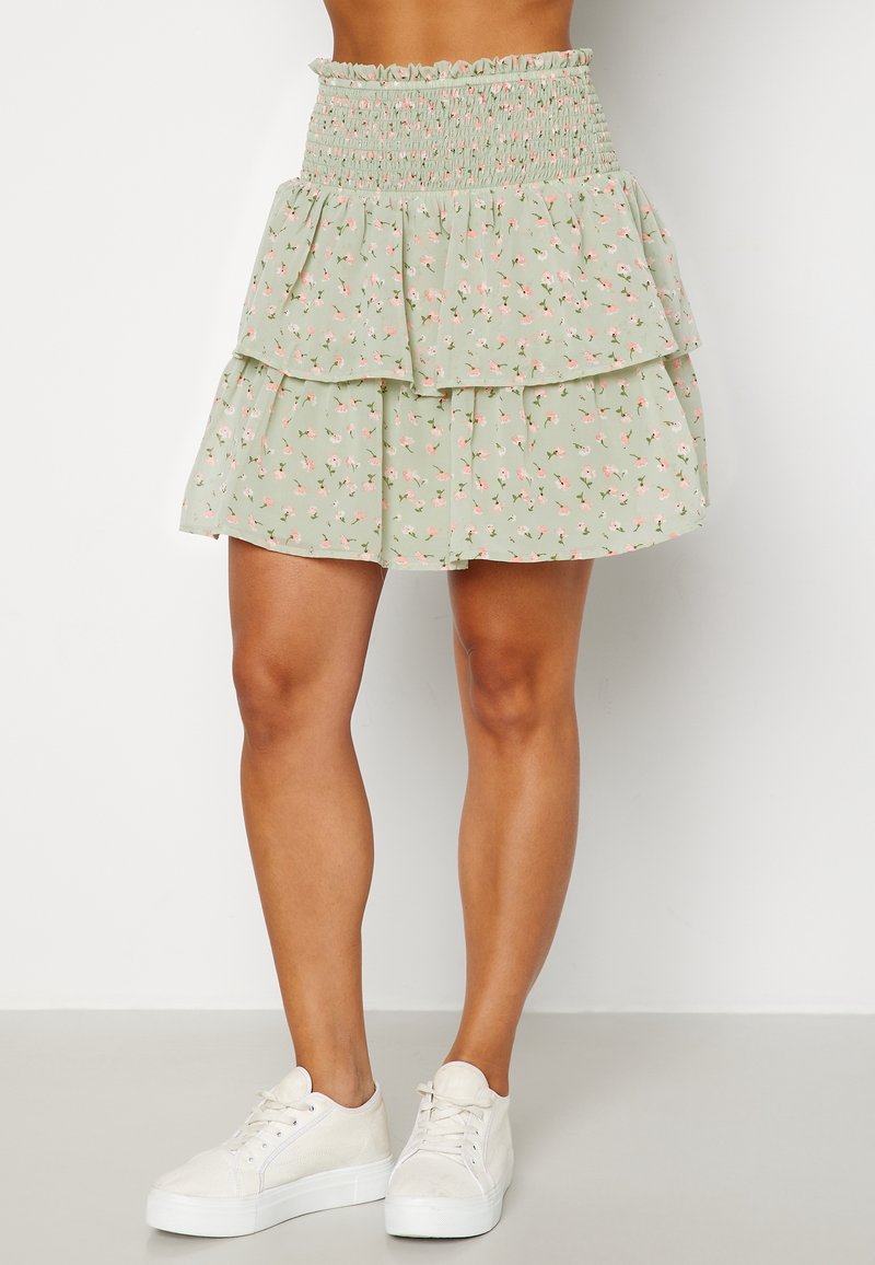 Falda verde de capas con patrón floral, cintura fruncida, tejido ligero, hasta la rodilla, combinada con zapatillas blancas.