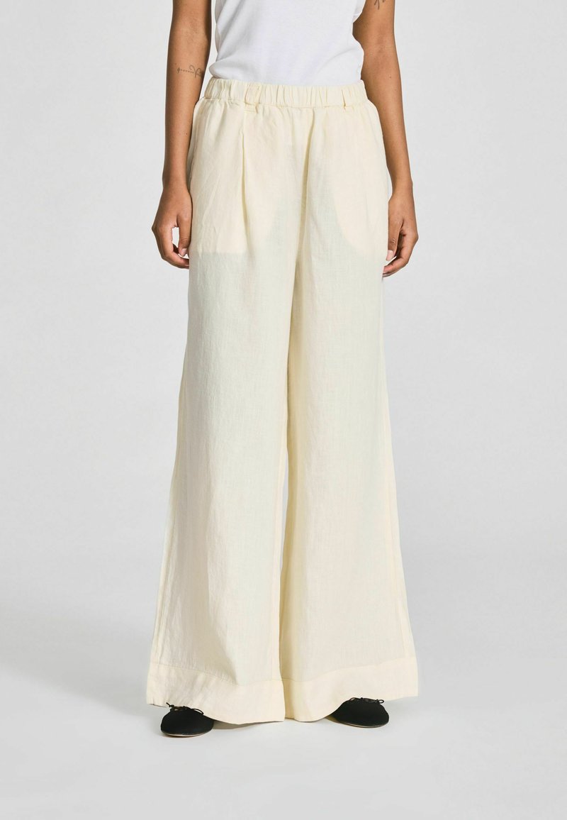 Personne portant un pantalon ample, taille haute, à jambes larges de couleur crème avec une ceinture élastique et des chaussures noires, debout devant un fond uni.