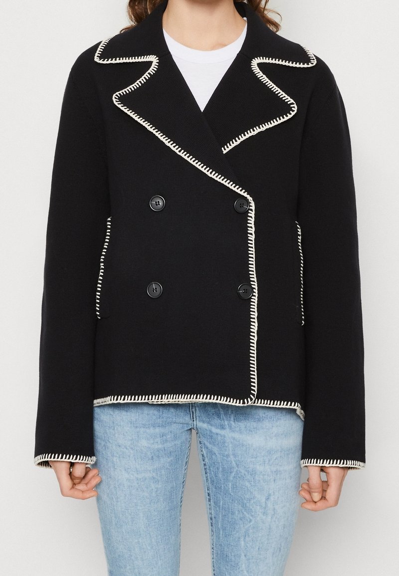 Veste noire à double boutonnage avec bordure en point de fouet blanche, larges revers et poches avant, portée sur une chemise blanche et un jean bleu clair.