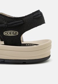 Sorte stofsandaler med en tekstureret rem, beige mellemsål og sort ydersål. KEEN-logoet er synligt på remmen. Moderne, ergonomisk design.
