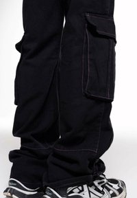 Un pantalon cargo noir présente de multiples grandes poches, des coutures contrastantes violettes et une coupe décontractée, associé à des chaussures de sport noir et blanc.