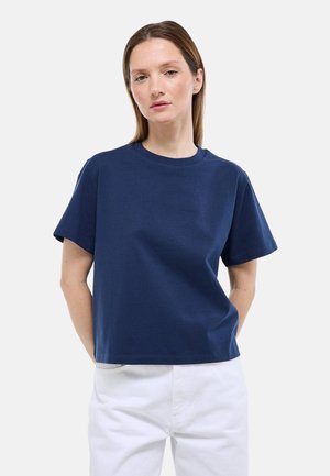 Femme portant un t-shirt bleu marine à manches courtes et un pantalon blanc, debout les mains dans le dos sur un fond clair uni.