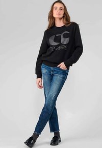 Sweat-shirt noir avec un logo brillant, associé à un jean bleu clair déchiré. Le mannequin porte des chaussures foncées et des chaussettes grises. Fond simple.