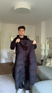 Svart polstret vest med høy krage, med glidelås foran og sidelommer, holdt av en person i innendørs miljø.