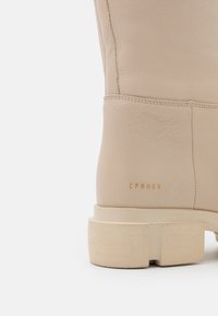 Bottine en cuir beige avec un talon bloc et un marquage en relief "CPNHGN" sur le côté, dotée d'une texture lisse et de détails de couture minimaux.