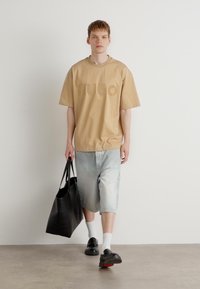 Tricou oversize bej cu logo embossat, pantaloni scurți rayati de un albastru deschis, sacoșa neagră și pantofi slip-on negri cu accente roșii.