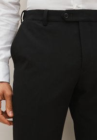Zwarte op maat gemaakte broek met een gladde textuur, voorzien van een platte voorkant, knoopsluiting en ceintuurlussen, gedeeltelijk getoond met een hand op de stof.