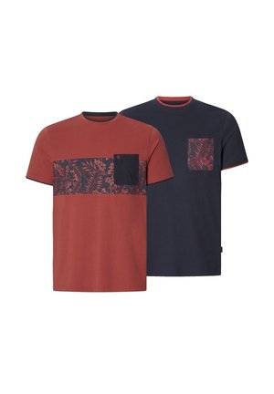 Mann mit tätowiertem Arm trägt ein rostfarbenes T-Shirt mit Blumenmuster und navyfarbener Tasche, kombiniert mit blauen Jeans, steht vor weißem Hintergrund.