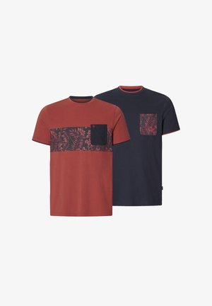 Mann mit tätowiertem Arm trägt ein rostfarbenes T-Shirt mit Blumenmuster und navyfarbener Tasche, kombiniert mit blauen Jeans, steht vor weißem Hintergrund.