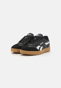 Tenis pretos em materiais de camurça e borracha, com detalhes brancos e sola de borracha. Design clássico de cano baixo com atacadores e língua com marca.