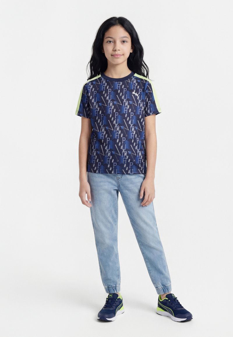 Fille aux longs cheveux noirs porte une chemise bleu marine à motifs, un jean bleu clair et des baskets bleu marine avec des accents vert néon, debout sur fond blanc.