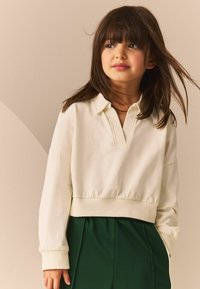 Cremefarvet pullover med polo krave, elastisk nederste kant og lange ærmer, parret med mørkegrønne højhvide bukser; blød tekstur.