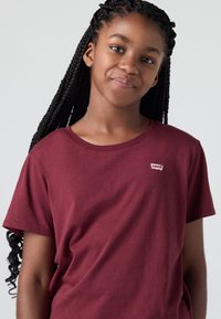 Jauna meitene ar garām pītām matiem, ģērbta bordo krāsas Levi's t-kreklā, maigi smaida pret vienkāršu gaišu fonu.