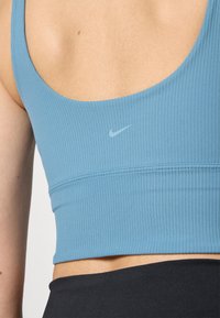 Μπλε ριγέ αθλητικό σουτιέν με σχήμα σκαφτού πίσω μέρους, που διαθέτει ένα διακριτικό λογότυπο της Nike σε ανοιχτό γκρι πάνω από τη ραφή. Μαλακή υφή και εφαρμοστό σχέδιο.