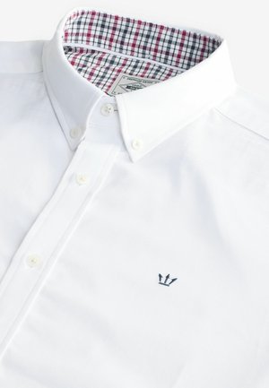 Camicia bianca in cotone con colletto abbottonato, caratterizzata da un motivo a scacchi nella fodera del collo e un piccolo ricamo di una corona blu marina sul petto.