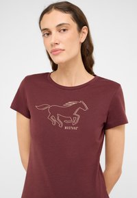 Camiseta de manga corta burdeos con un contorno de un caballo corriendo en color dorado. Texto "MUSTANG" mostrado debajo del gráfico. Textura suave.