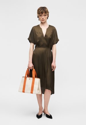 Γυναίκα με μεσοφόρεμα ελιάς κρατώντας λευκή τσάντα ώμου με πορτοκαλί χερούλια, φορούντας μαύρες μυτερές γόβες, στέκεται μπροστά σε απλό φόντο.