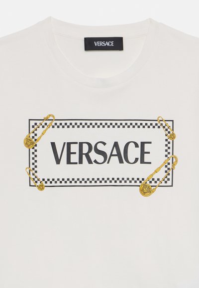 Vit bomull t-shirt med en svart "VERSACE" logo inom en rektangulär ram och detaljer av guld säkerhetsnålar.
