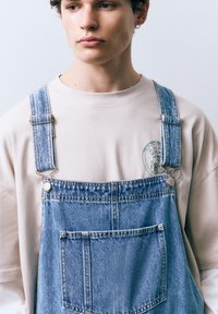 Spijkerbroek-overalls met verstelbare bandjes, lichtblauwe kleur, voorzak, gedragen over een beige T-shirt met een groen grafisch ontwerp.
