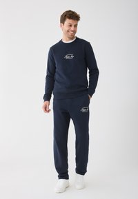 Marineblauwe sweatshirt en bijpassende broek van zachte stof, met een logo op de borst en zijzakken, gecombineerd met witte sneakers.