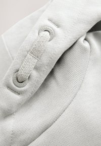 Tela gris claro con una textura suave. Presenta un botón y una correa tejida, mostrando costuras detalladas y la marca en el botón.