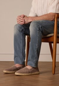 Espadrillas in suede marrone con suole in corda di juta; indossate con jeans di denim azzurro chiaro. L'ambientazione include una sedia di legno e uno sfondo semplice.