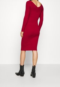 Robe tricotée rouge côtelée avec un dos en V et des manches longues, silhouette ajustée, associée à des bottines noires à la cheville, se tenant sur un fond blanc.