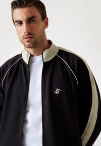 Veste zippée noire avec un col montant, des accents beiges contrastés et un logo sur le côté gauche. Portée sur une chemise blanche.