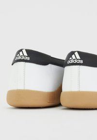 Tenis brancos com detalhes pretos e três riscas brancas no calcanhar, apresentando uma sola de borracha e parte superior de sintético liso.