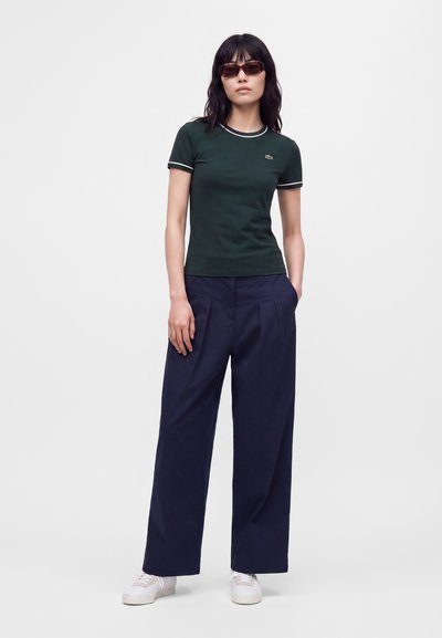 Femme portant un t-shirt ajusté manches courtes vert foncé, pantalon large bleu foncé, baskets blanches et lunettes de soleil, debout avec une main dans la poche.